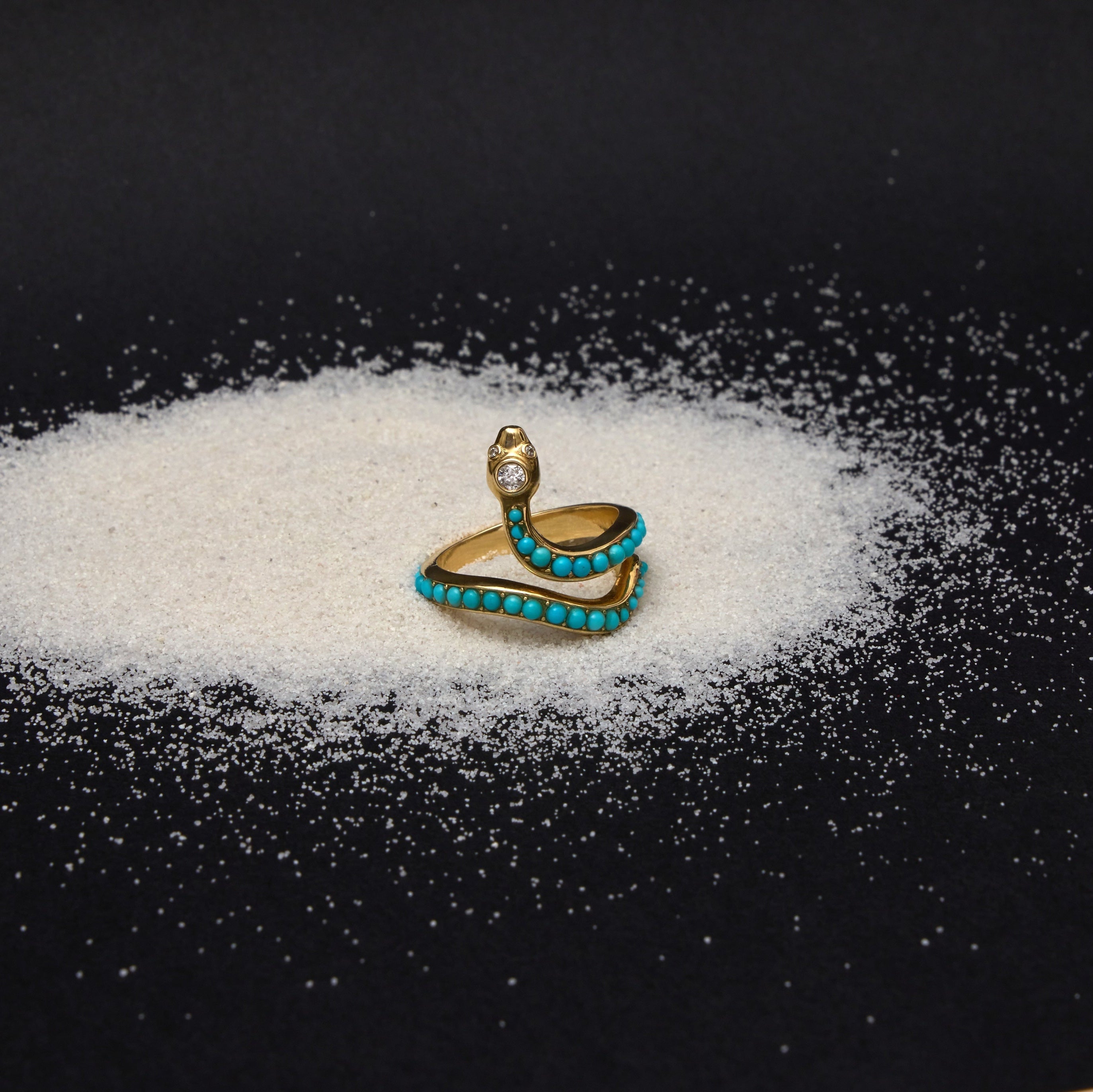 A gold snake ring with turquoise blue stones, positioned on top of a small pile of white sugar on a black background.