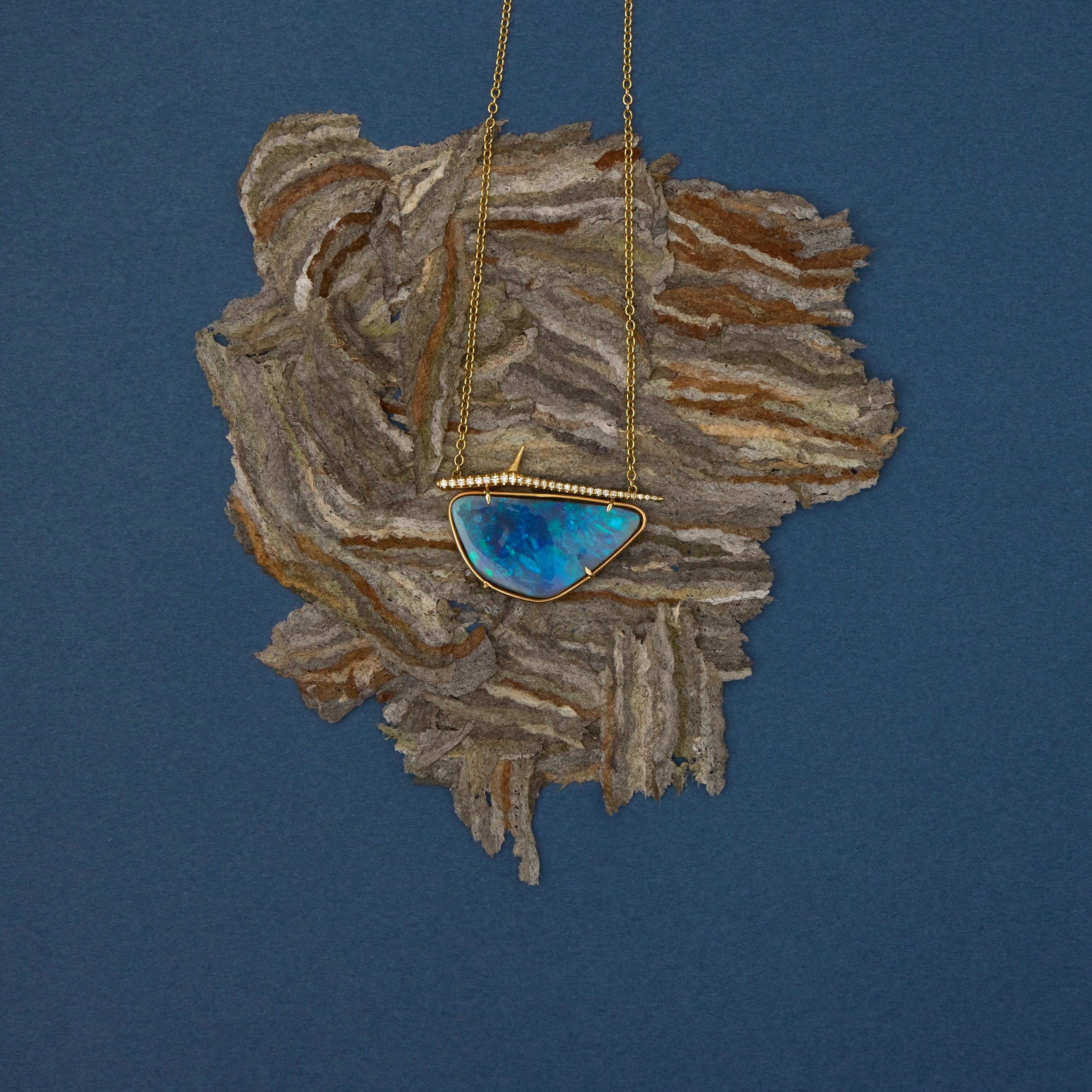 A gold necklace with a blue-green iridescent stone pendant, displayed on a piece of textured driftwood against a solid dark blue background.