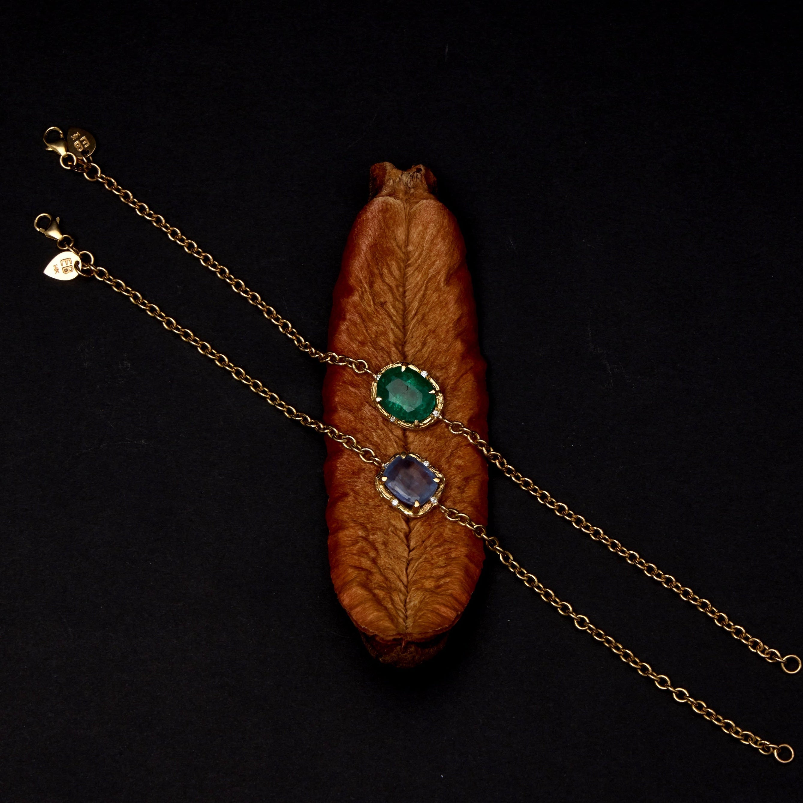 A gold chain bracelet with two oval gemstone settings, one green and one blue, displayed on a dark background with a decorative wooden leaf prop.
