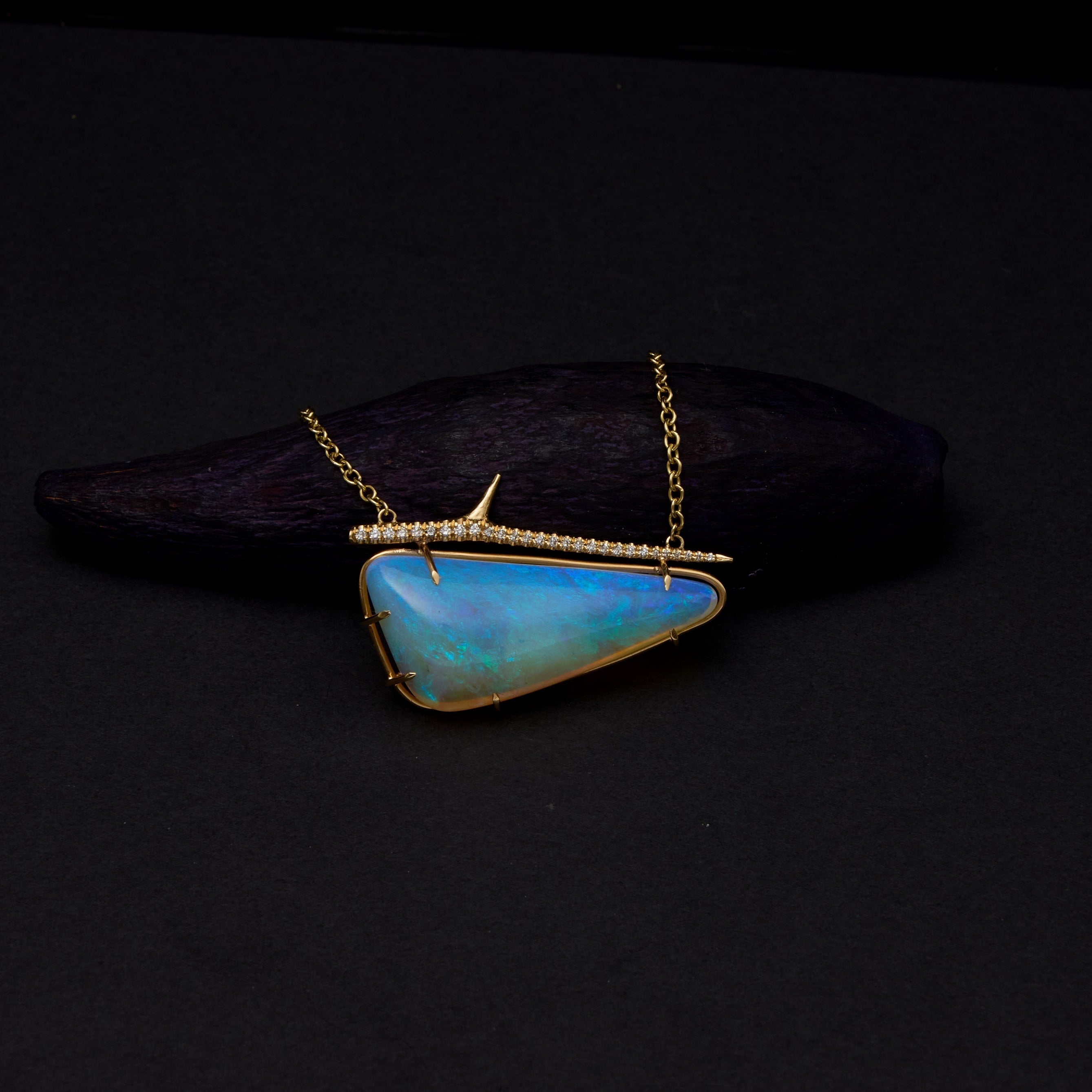 A gold necklace with a triangular blue-green opal pendant, displayed on a dark wooden block against a black background.