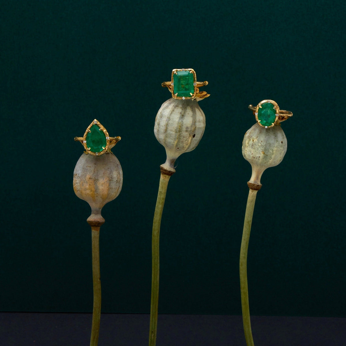 Three vintage gold rings with green gemstones mounted on artificial poppy pods, displayed on metal stands against a dark green background.