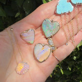 A hand holding several iridescent heart-shaped and shell-shaped pendant necklaces with gold-tone chains, photographed outdoors against green foliage.
