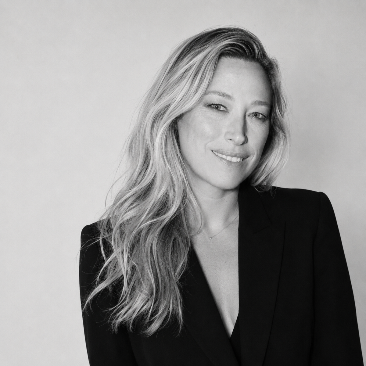 A black and white portrait of a smiling woman with long wavy hair, wearing a dark blazer against a plain light background.