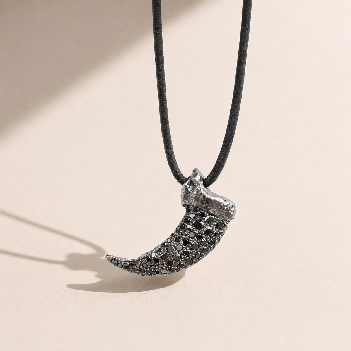 A silver crescent moon pendant covered in small dark pavé stones, hanging from a black cord necklace against a warm beige background with soft shadows.