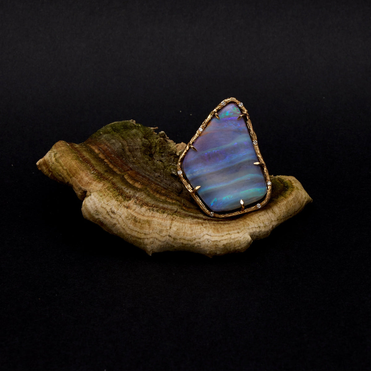 A raw, irregularly shaped blue opal gemstone with iridescent blue and purple shifts, set in a gold bezel and displayed on a piece of natural wood against a solid black background.