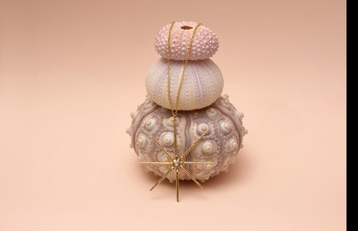 A gold-toned starburst pendant necklace with a central clear stone, draped over a stacked set of three sea urchin shells in pastel pink, cream, and lavender on a solid peach background.