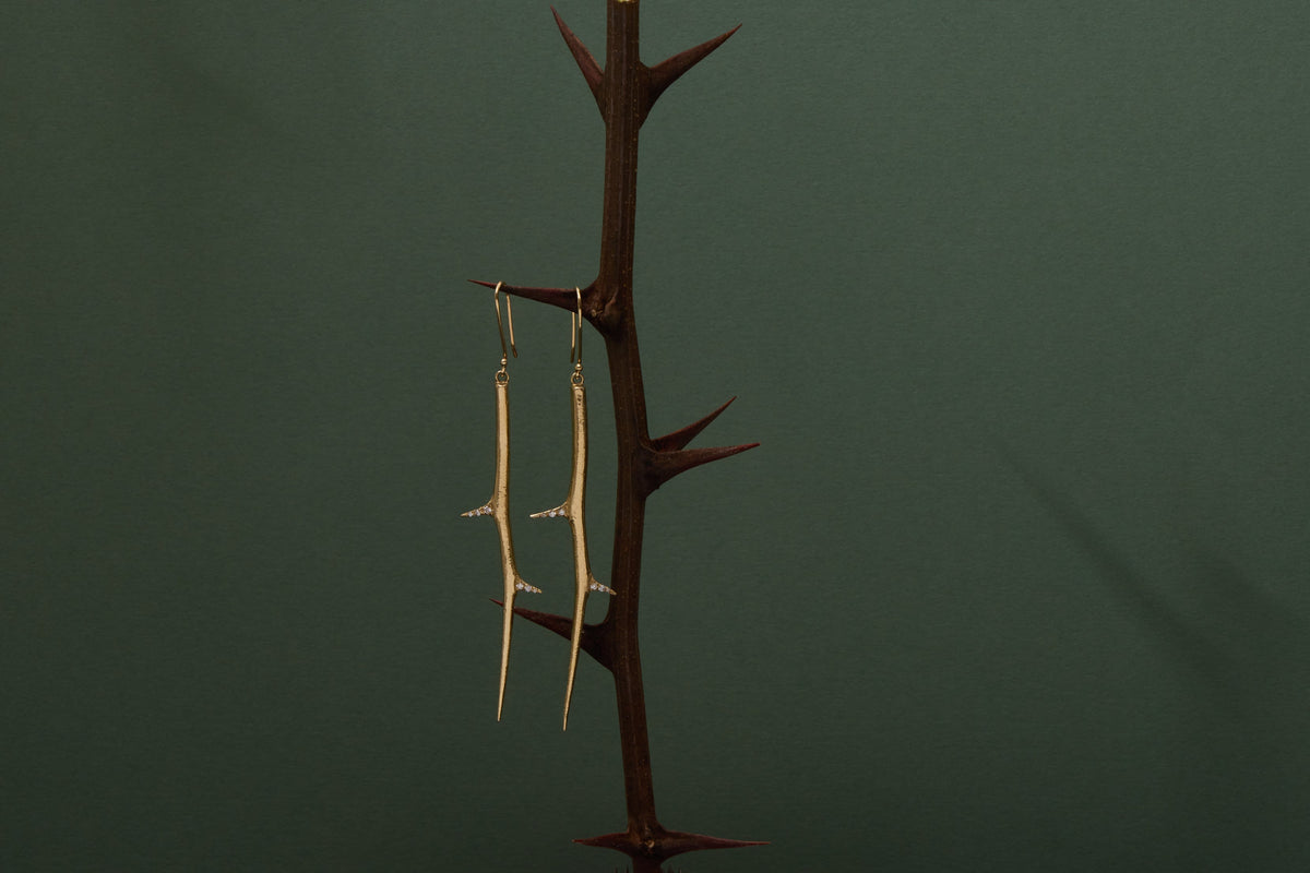 A pair of long, thin gold-toned earrings displayed on a dark brown thorn stand, which is placed on a pink block against a dark green background.