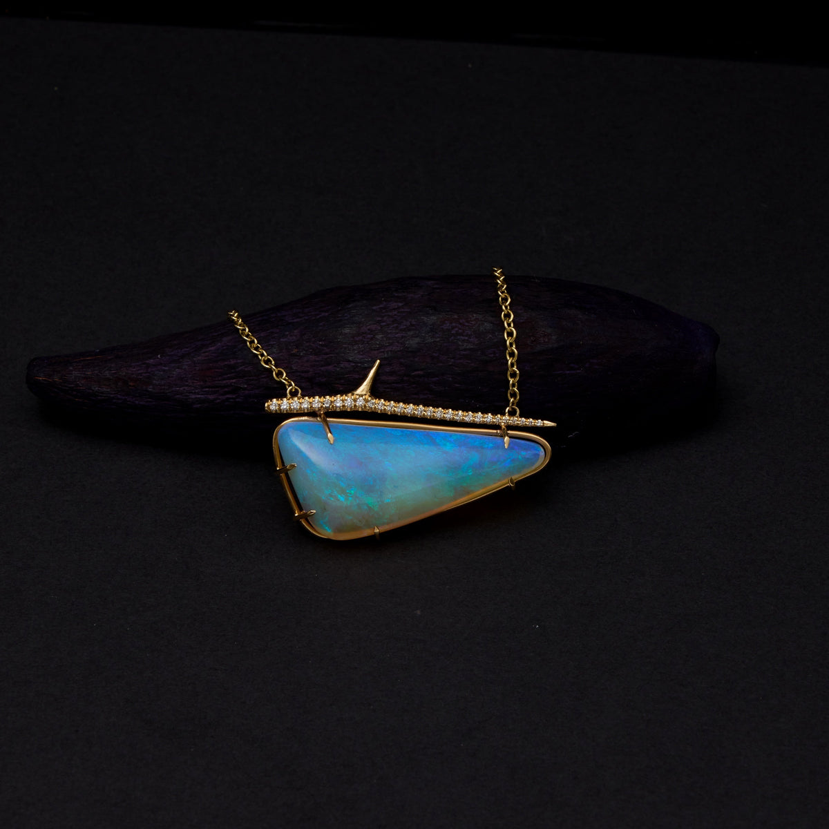 A gold necklace with a triangular blue-green opal pendant, displayed on a dark wooden block against a black background.