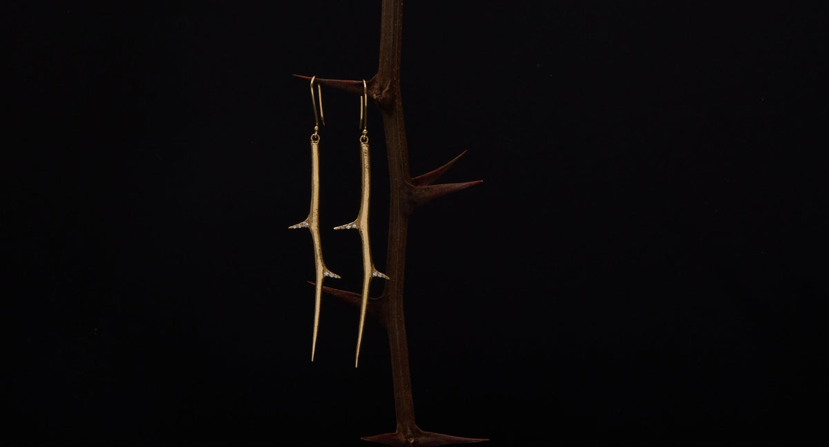A pair of long, slender gold-toned earrings hanging from a decorative branch-shaped display stand on a pink pedestal against a black background.
